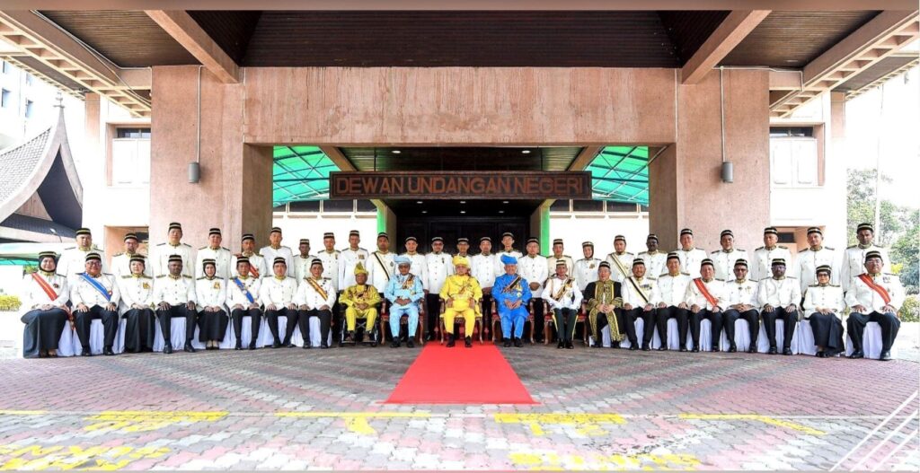 Group of Formally Dressed Officials in White Uniforms Posing for a Ceremony on a Red Carpet in Front of a Government Building, with Central Dignitaries in Bright Traditional Attire Beneath a Signboard.**oops**