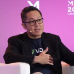 Speaker Wearing Glasses on a Pink Money20/20 Stage, Gestures with Hands While Seated in a Black Blazer in a Panel Discussion.